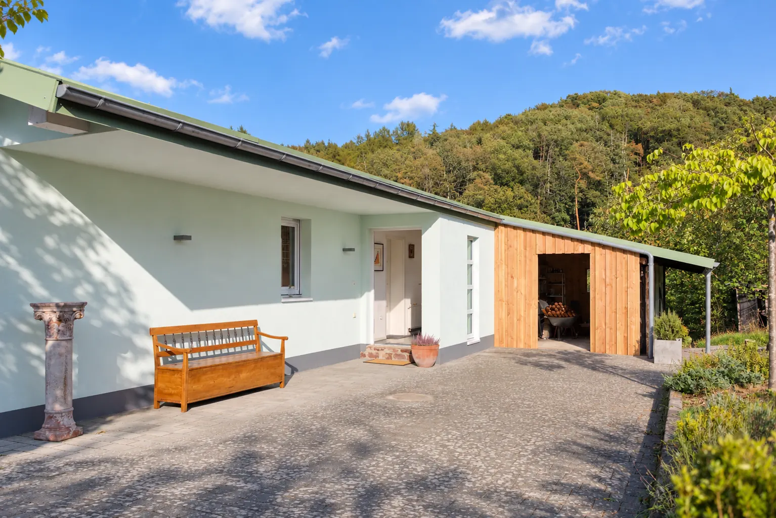 Das Ferienhaus von vorne mit Vorplatz, Holzbank und bewaldetem Hang dahinter